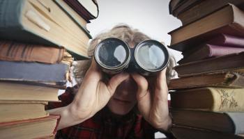 man using binoculars in between stack of books
